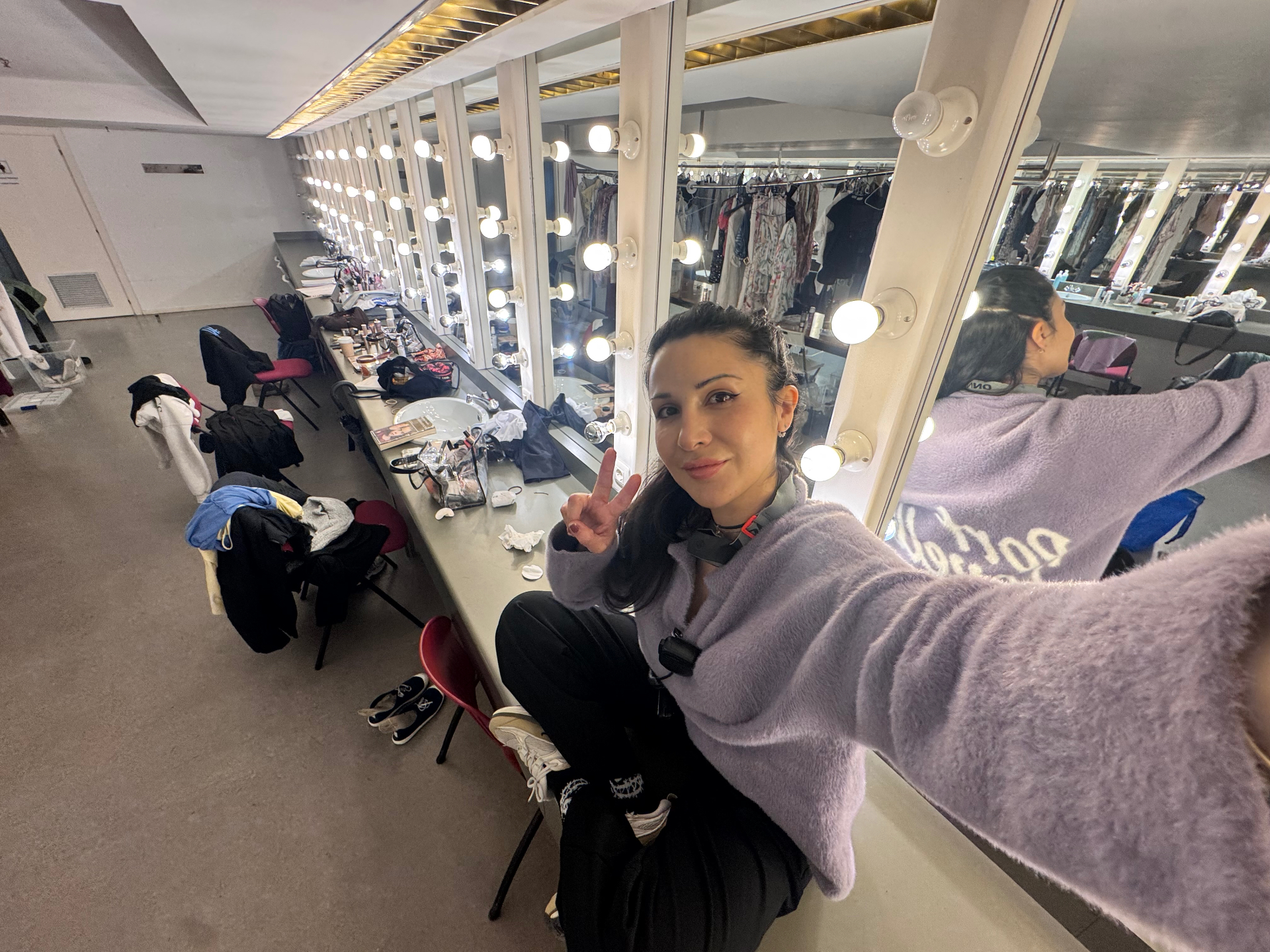 Maya in the theatre dressing room, wearing a lilac jumper among the costume rails